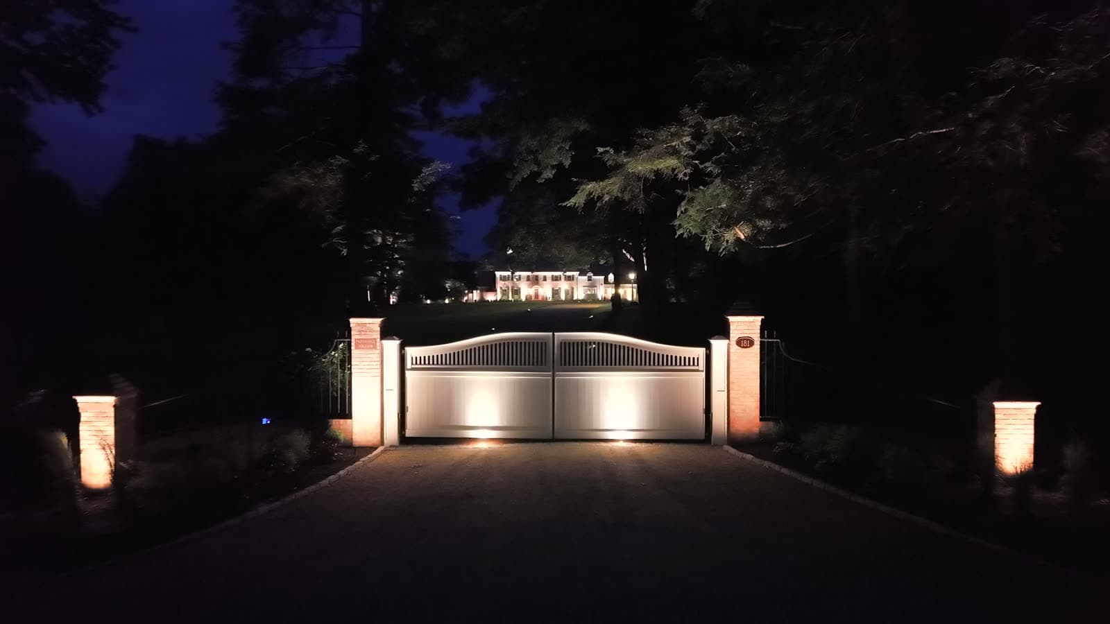 The lit gated entrance to a Farmington property at night, the house glowing at the top of the drive beyond